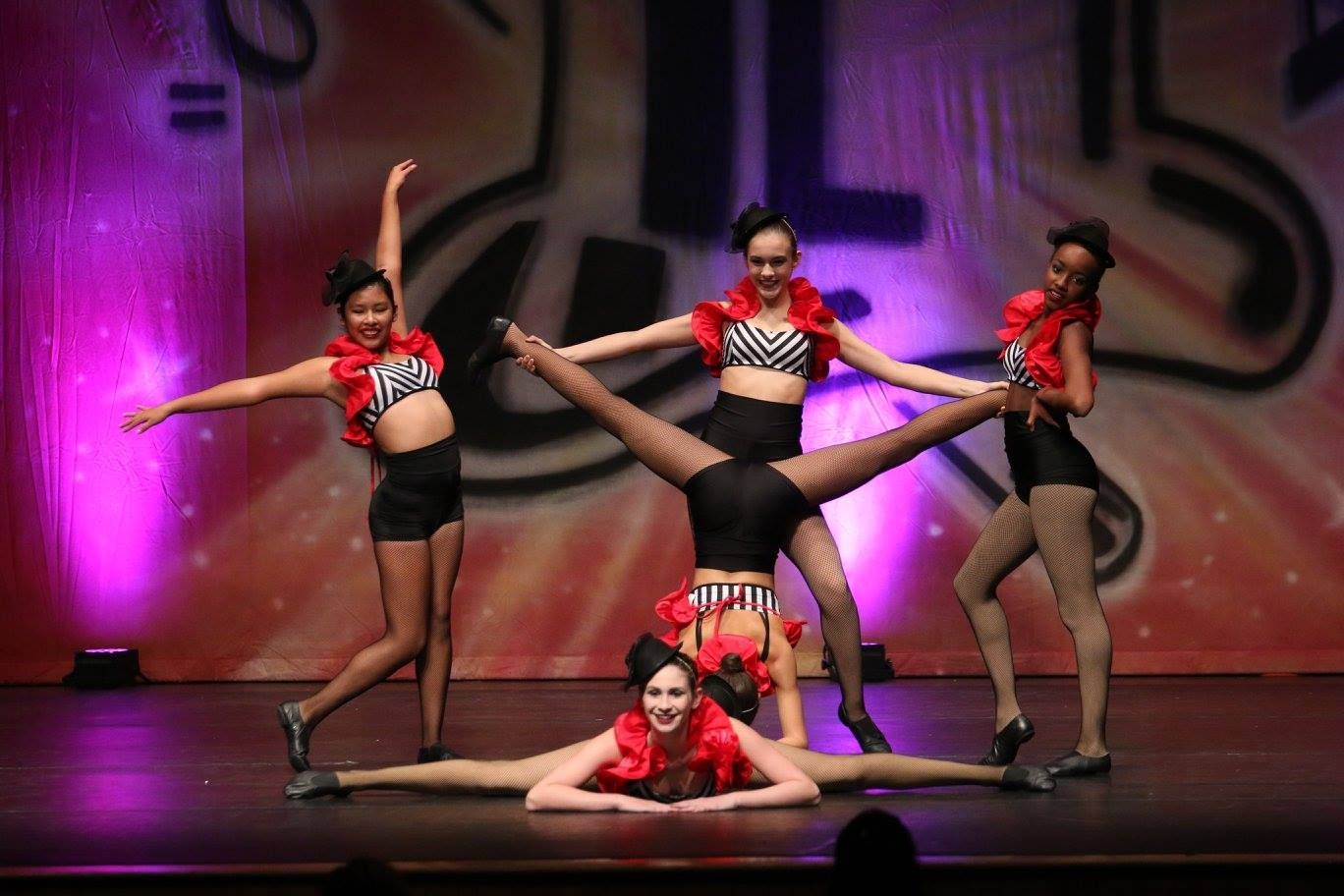 Five dancers in stage performance; acrobatic pose with split, leg splits, and red and black costumes.