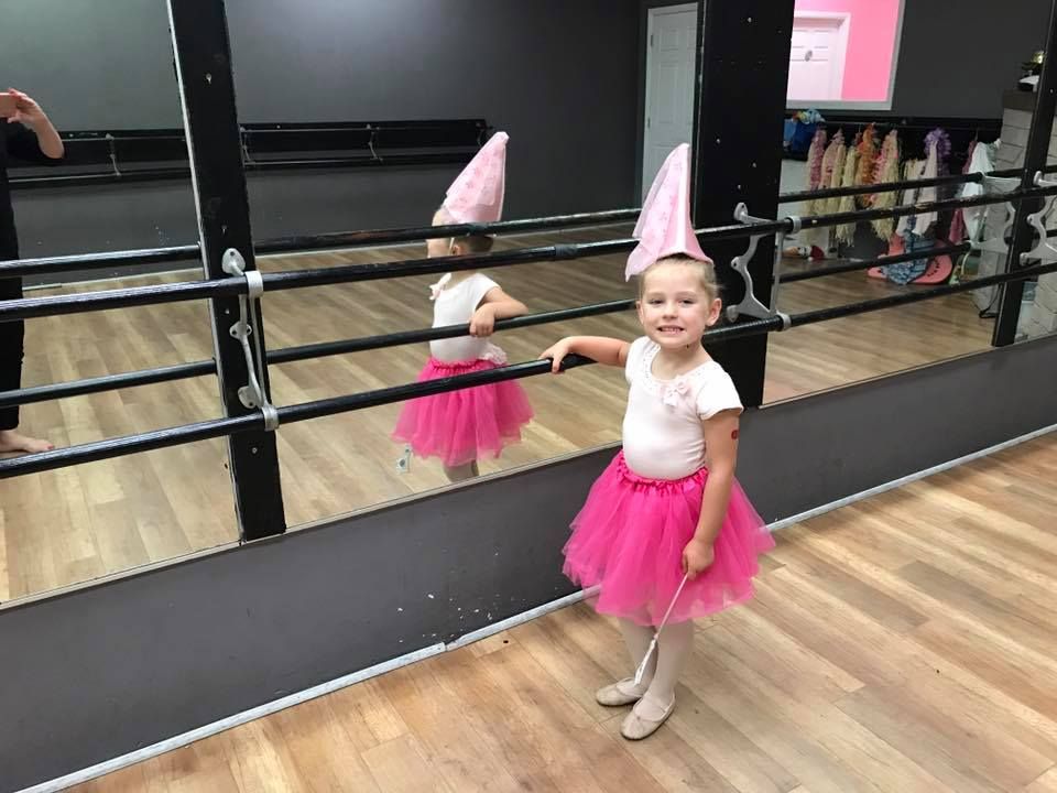Girl in pink tutu and hat, posing by a mirror in a dance studio, holding a wand.