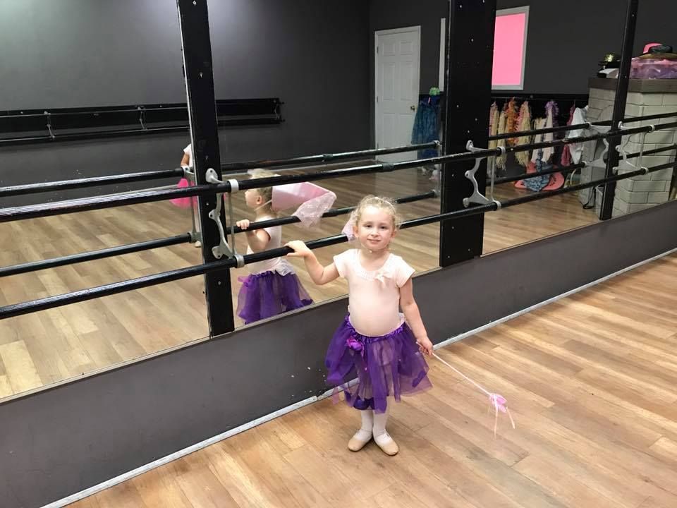 Young child in purple tutu and ballet slippers stands by dance studio mirror, holding a wand.