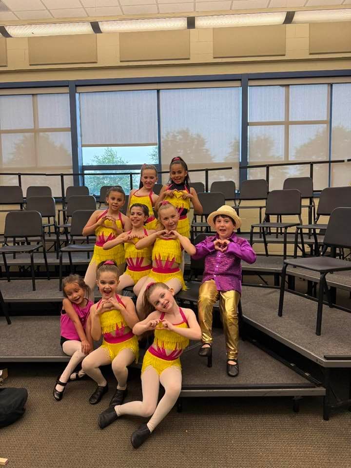 Group of children in dance costumes making heart shapes. They're on a stage, some with props.