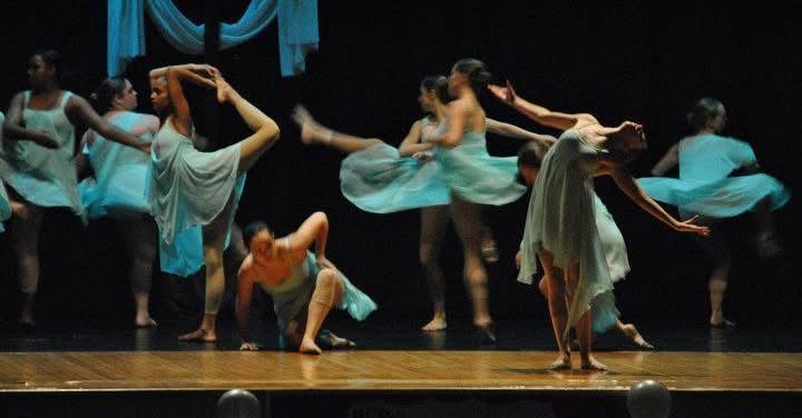 Dancers in flowing blue-green dresses perform on stage, illuminated by stage lights; some are in arabesque.