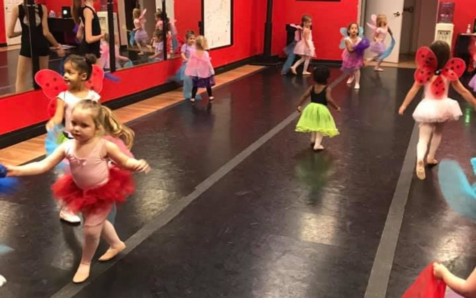 Young children in tutus and fairy wings dance in a studio with red walls and a mirror.