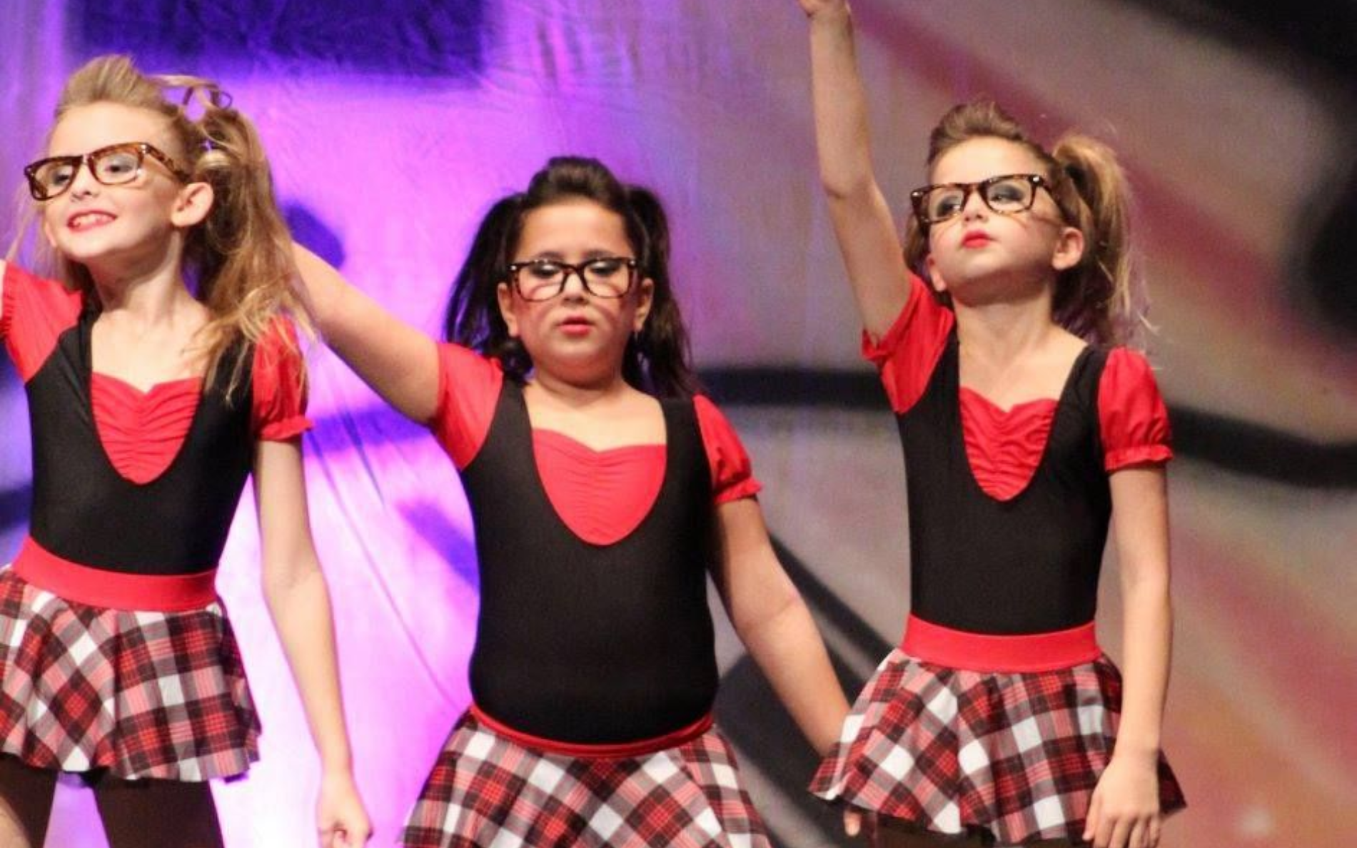 Three young dancers in black and red outfits with glasses, performing onstage.