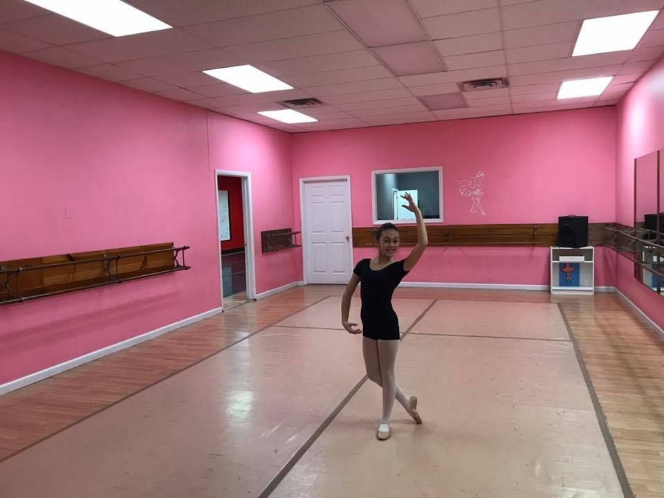 A person in ballet attire poses in a pink dance studio, arm raised, leg extended.