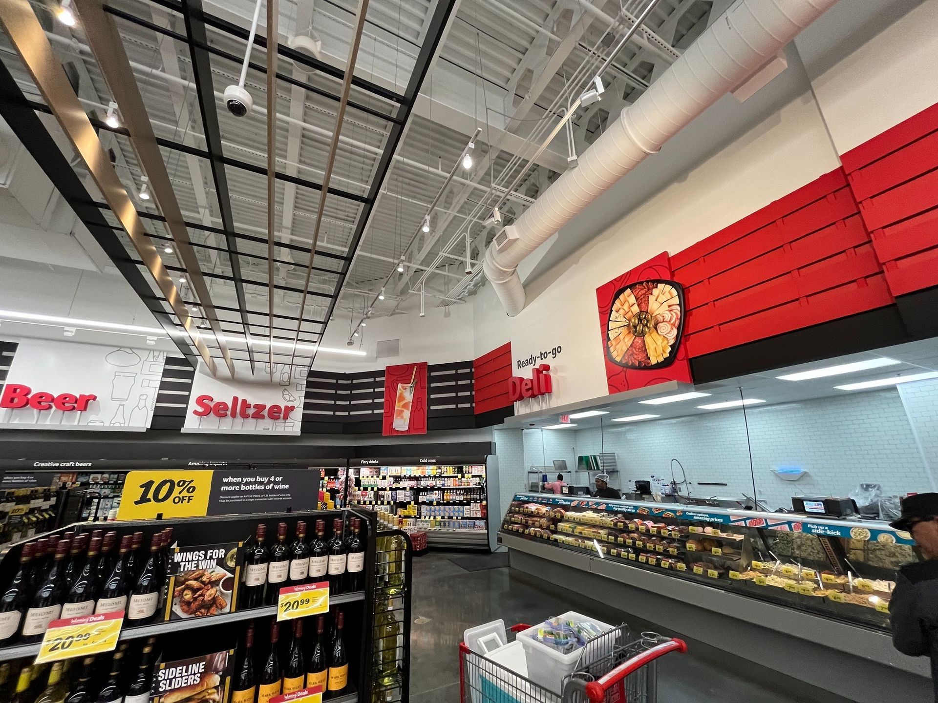 Wide supermarket aisle with refrigerated deli cases, red wall accents, and high industrial ceiling