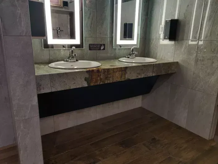 Modern public restroom with double sinks, lit mirrors, and a stone countertop over wood-look flooring