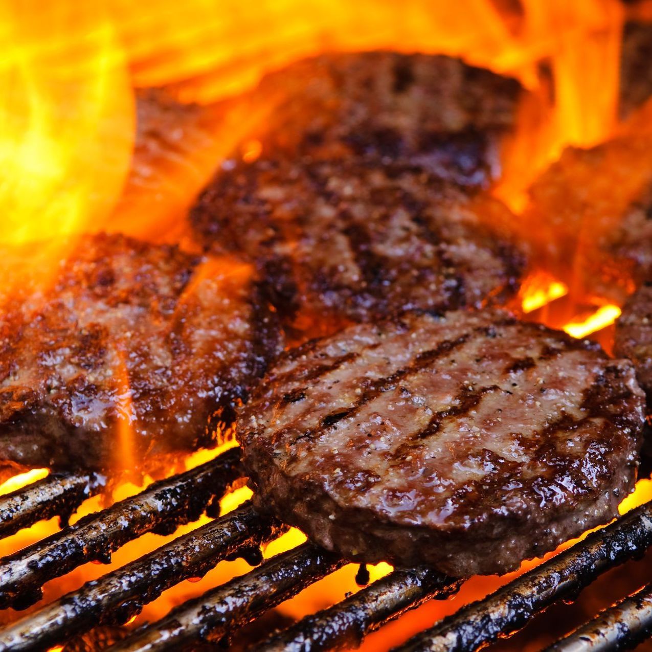 Hamburgers are cooking on a grill with flames in the background.