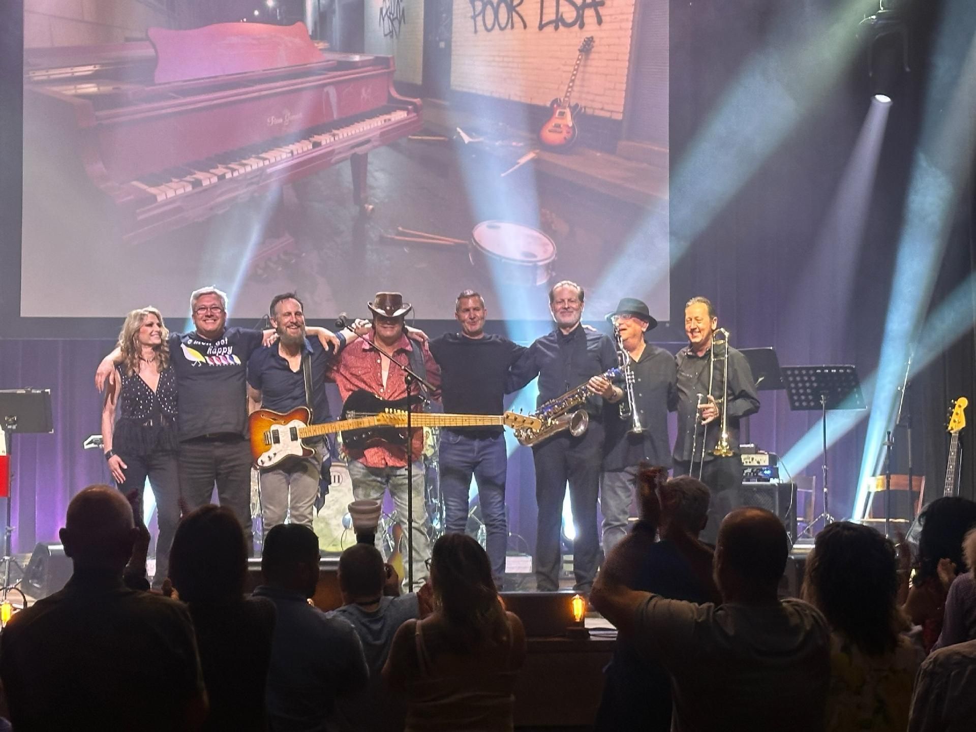 A band of eight musicians stands on a stage in front of an audience. A screen behind them displays a picture of a piano and the words 