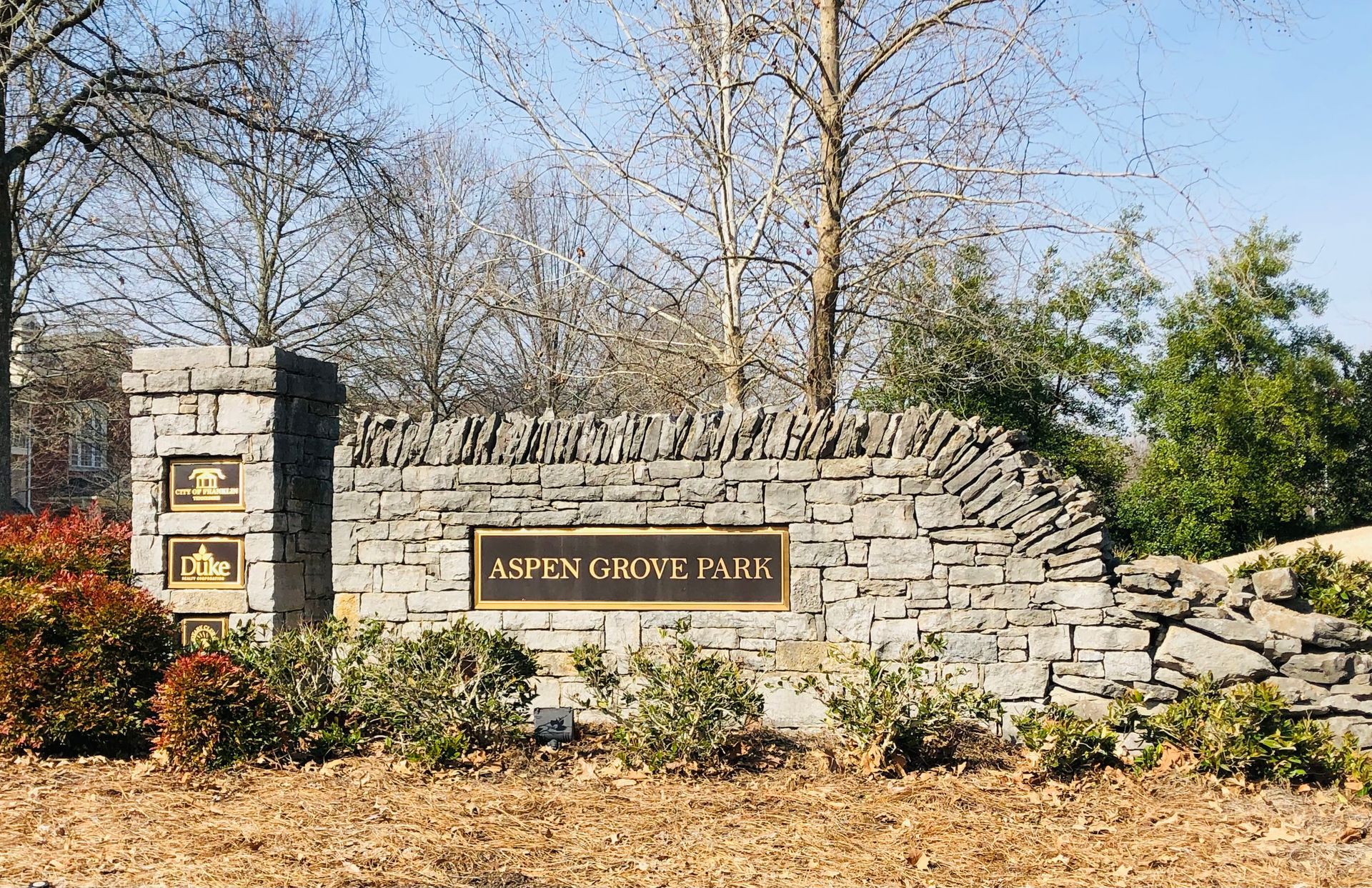 Stone sign for Aspen Grove Park with name plaque, bushes, and trees.