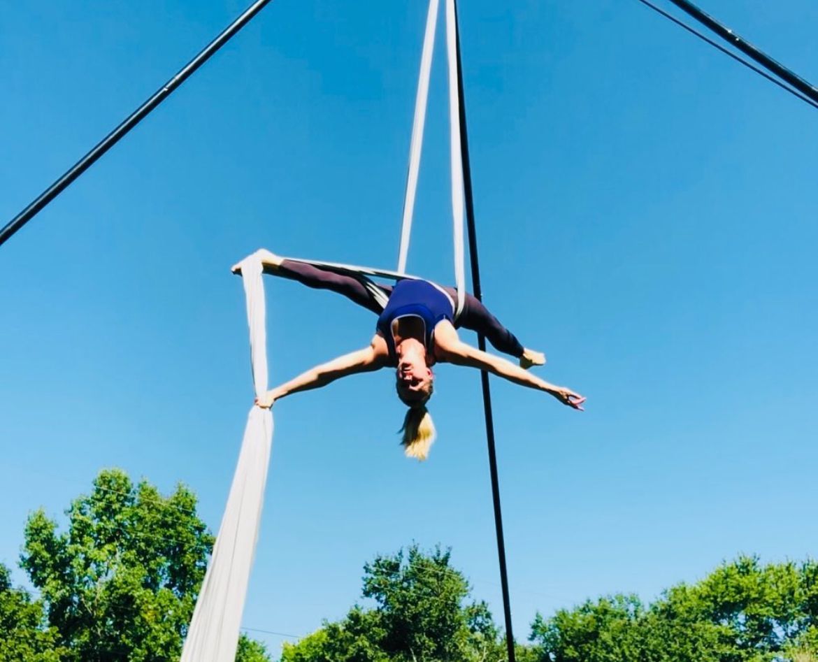 Woman performs aerial silks outdoors, splitting legs with arms outstretched, blue sky background.