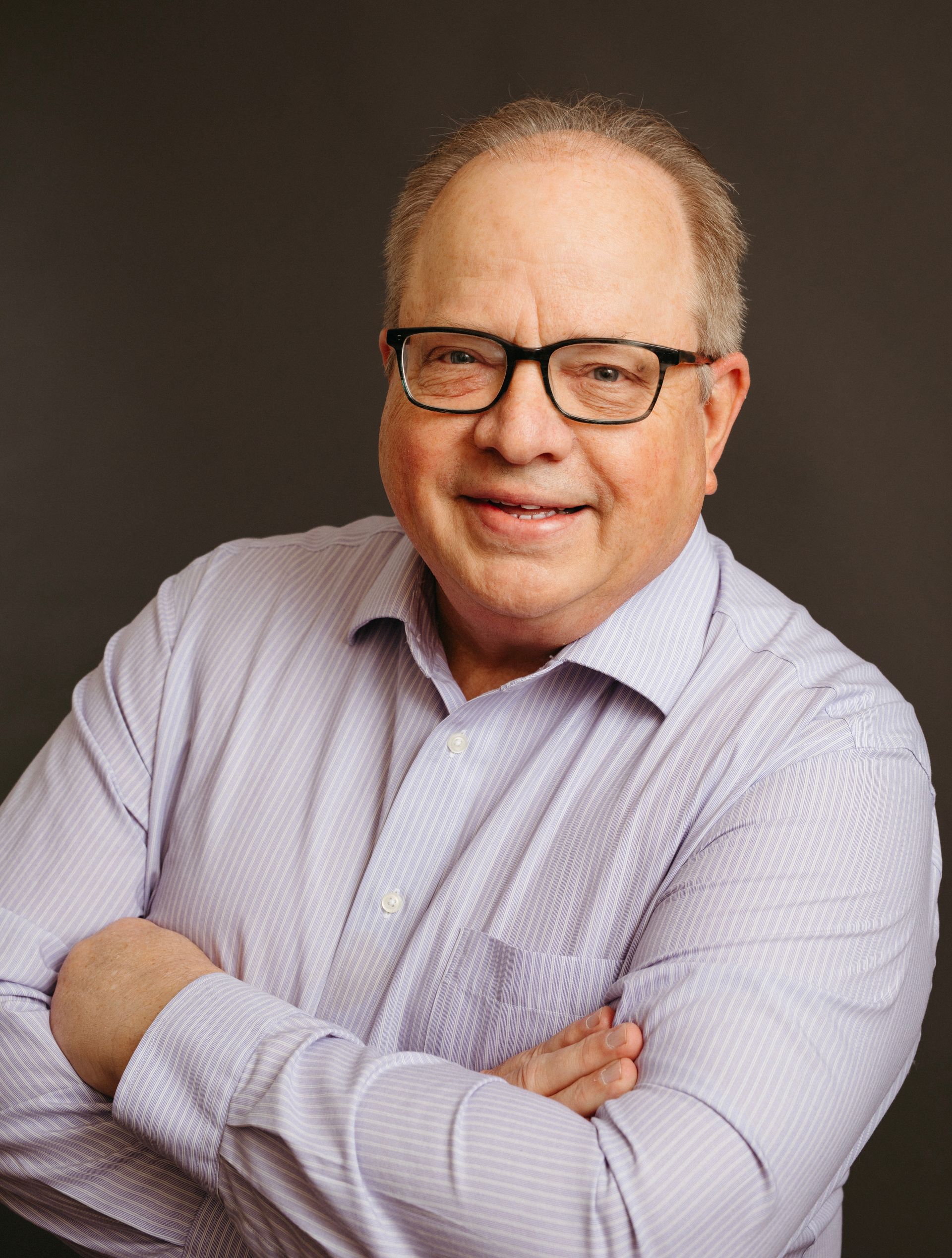 A man wearing glasses and a purple shirt is standing with his arms crossed.