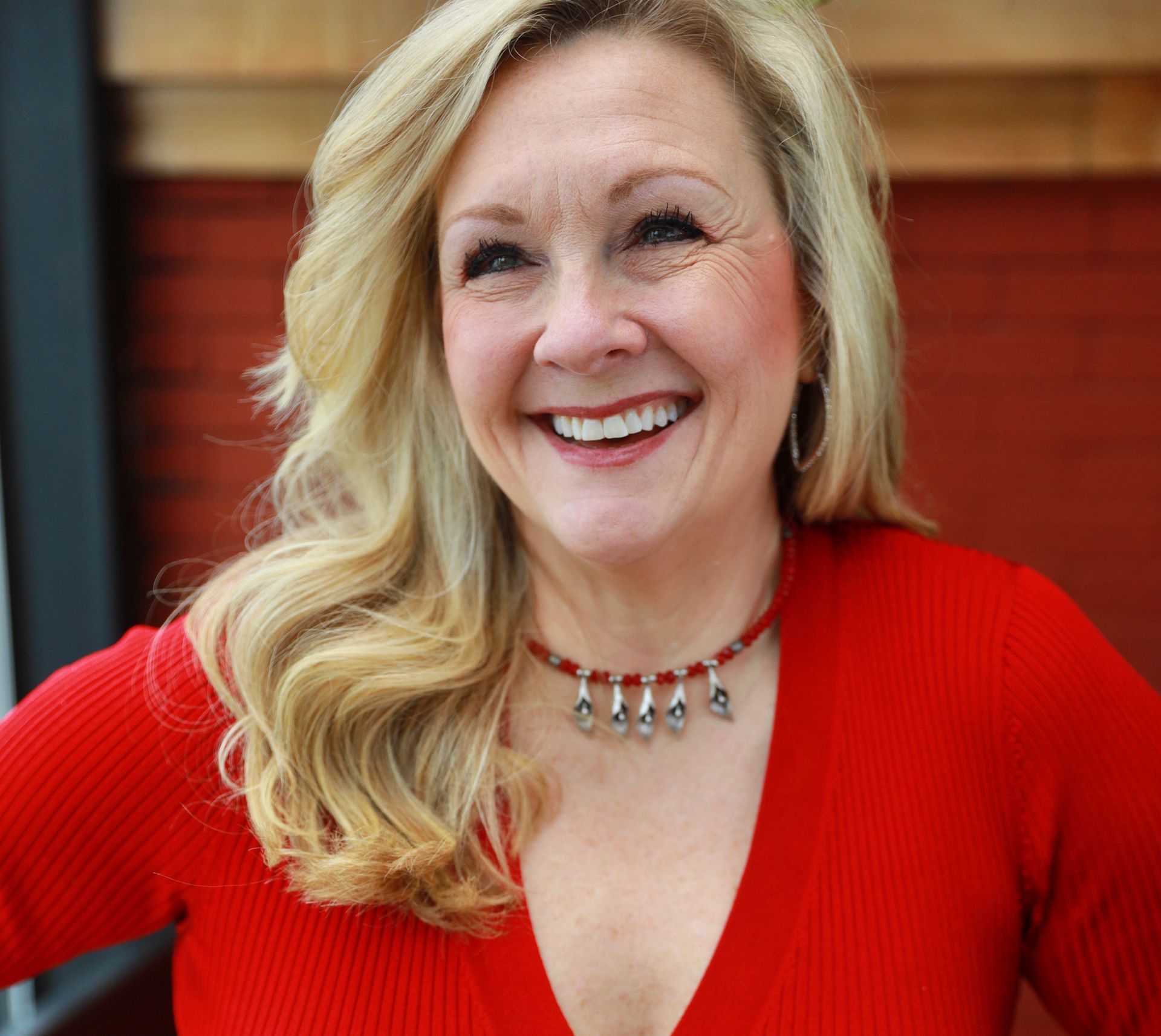 Blonde woman in red top smiling, wearing a red beaded necklace. Leaning against a brick wall.