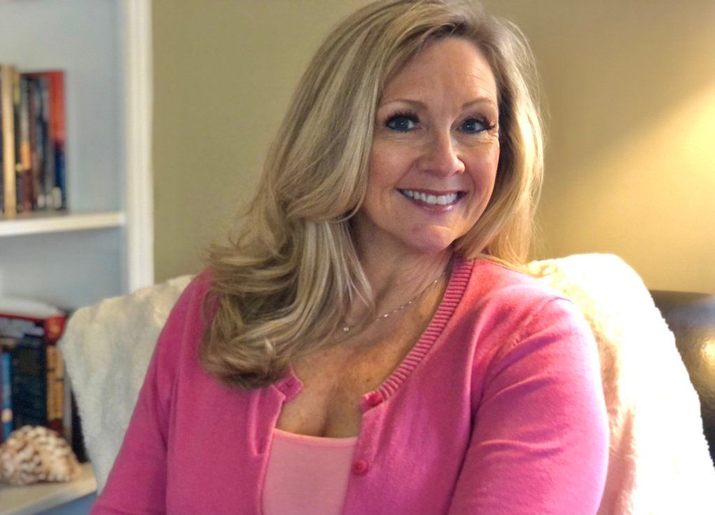 Woman with blonde hair smiles, wearing pink cardigan, sitting in a chair near a bookshelf.