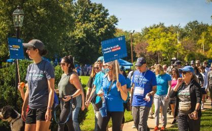 People walking outdoors at an event, many wearing blue shirts, some holding signs on a sunny day.