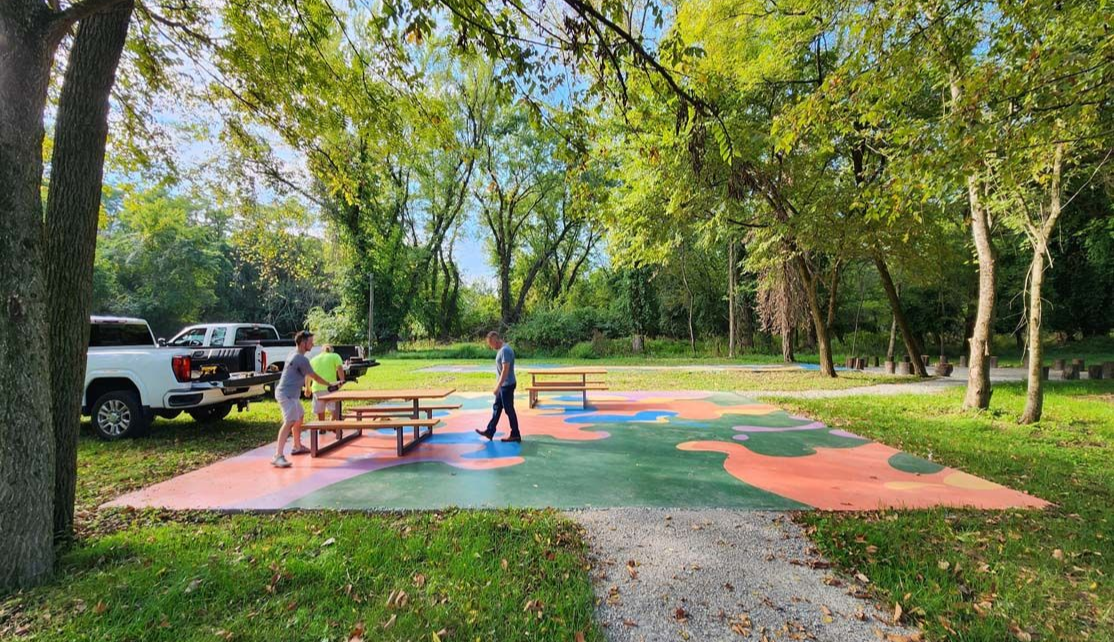 Two men setting up a picnic table on a colorful, paved area in a park with trees and trucks.