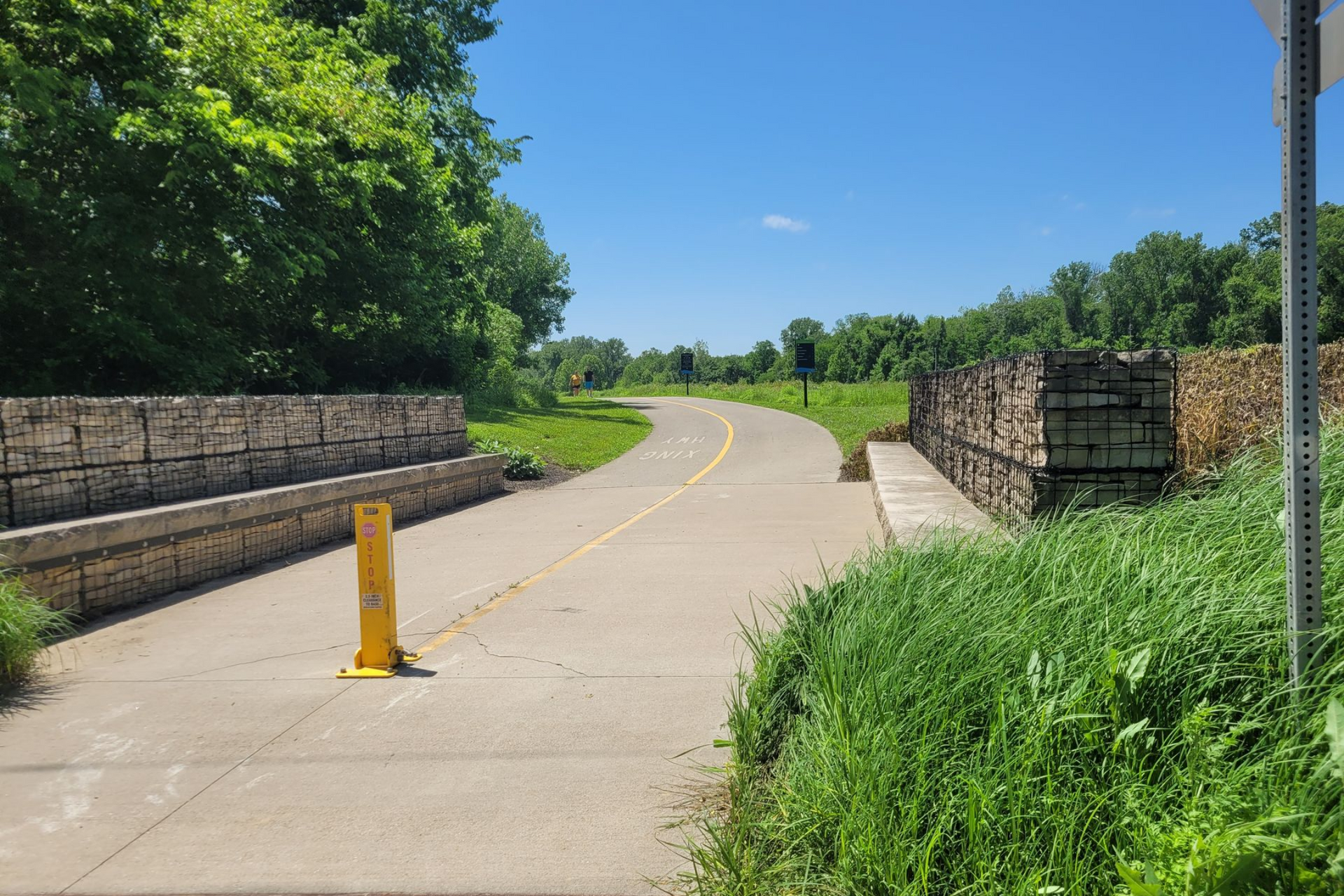 Paved path curves through green field, with stone walls on either side. Yellow pole blocks access; sunny day.