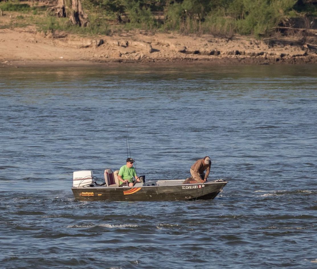 Two people on a small boat fishing in a river; one drives, the other leans over the side.