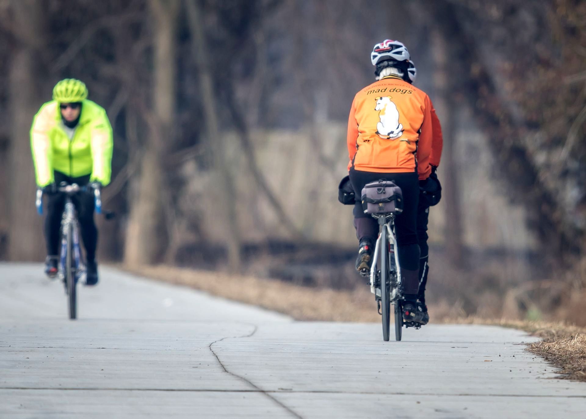 Two cyclists on the Ohio River greenway, one in yellow jacket, the other in orange jacket, trees in the background.