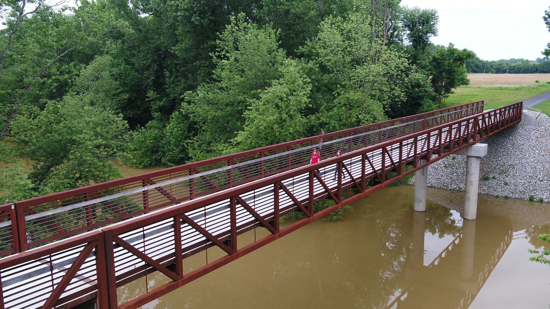 A person walks across a brown metal bridge over a muddy river, flanked by trees.