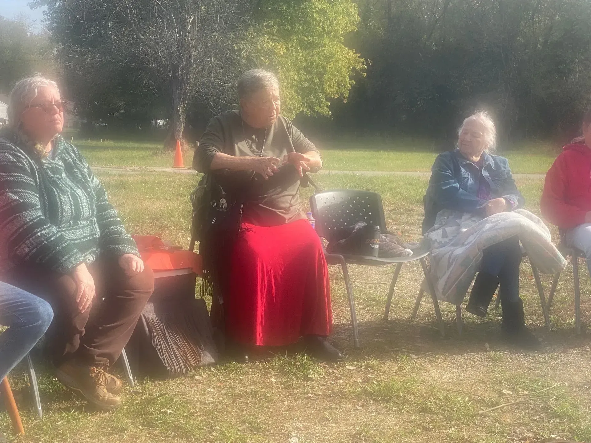 People seated in a circle outdoors; woman plays bagpipes, others listen; grassy field, sunny day.