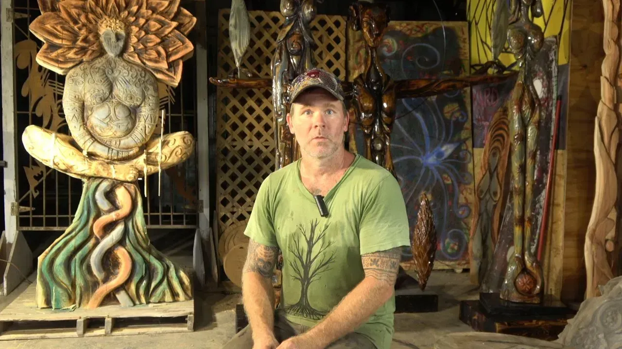 Man with tattoos sits in front of carved wooden figures. Setting is an art studio.