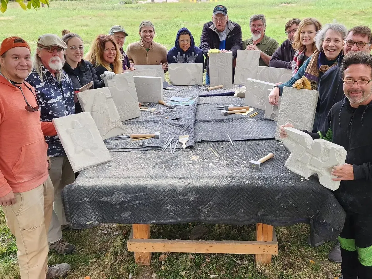A group of people carving stone outside, smiling. They stand around a table with tools and finished carvings.