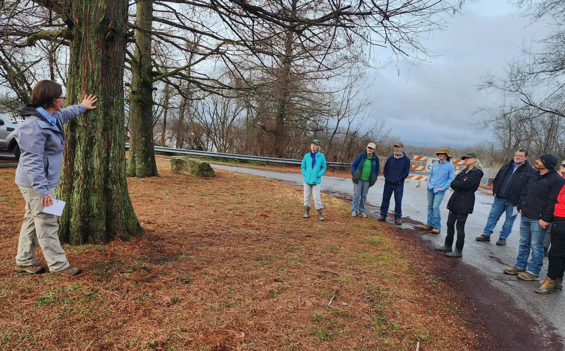 Woman gesturing to tree, leading a group on a nature walk along a paved path, fall foliage.