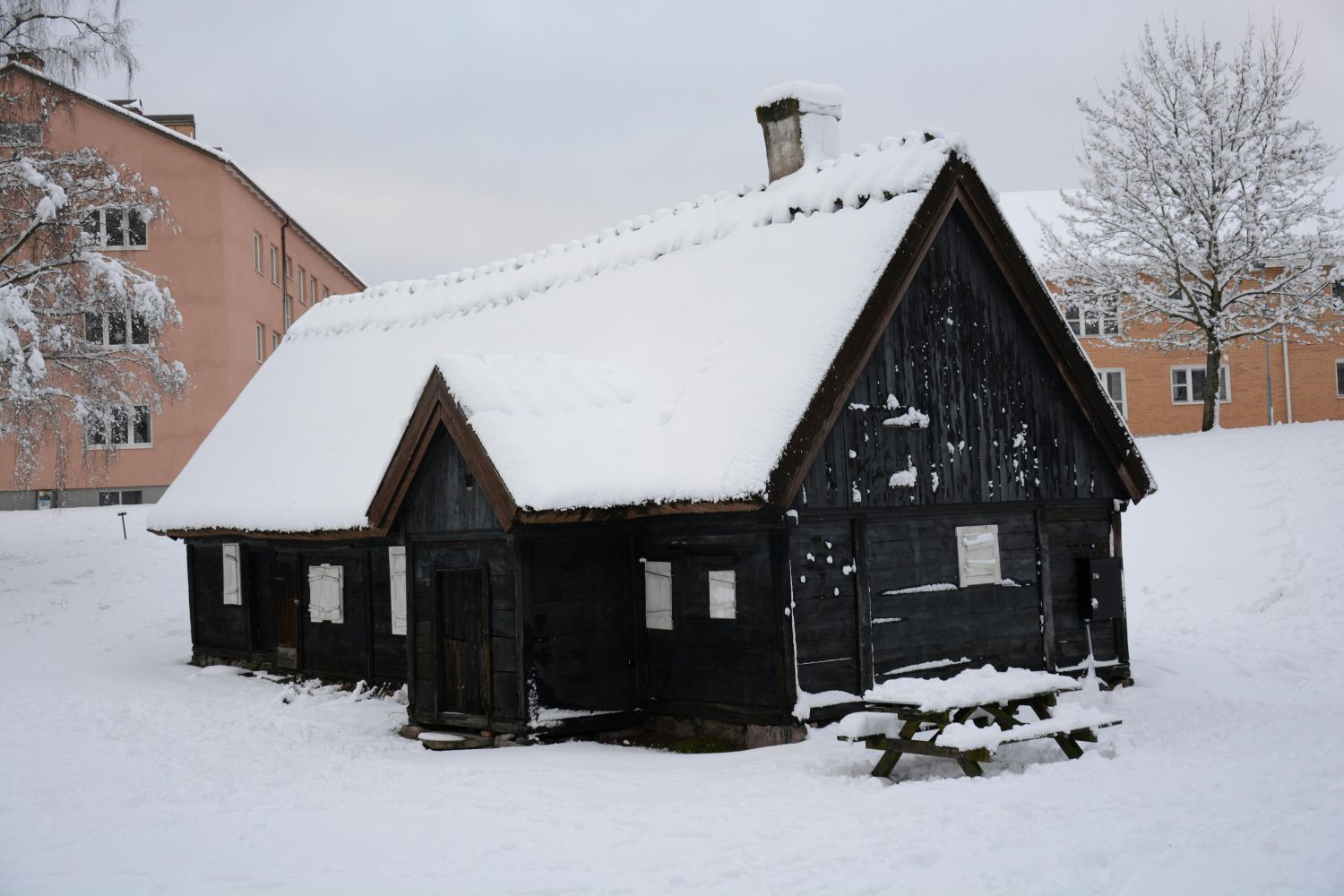 Snow-covered, dark wooden building with a peaked roof and chimney; a picnic table sits in front.