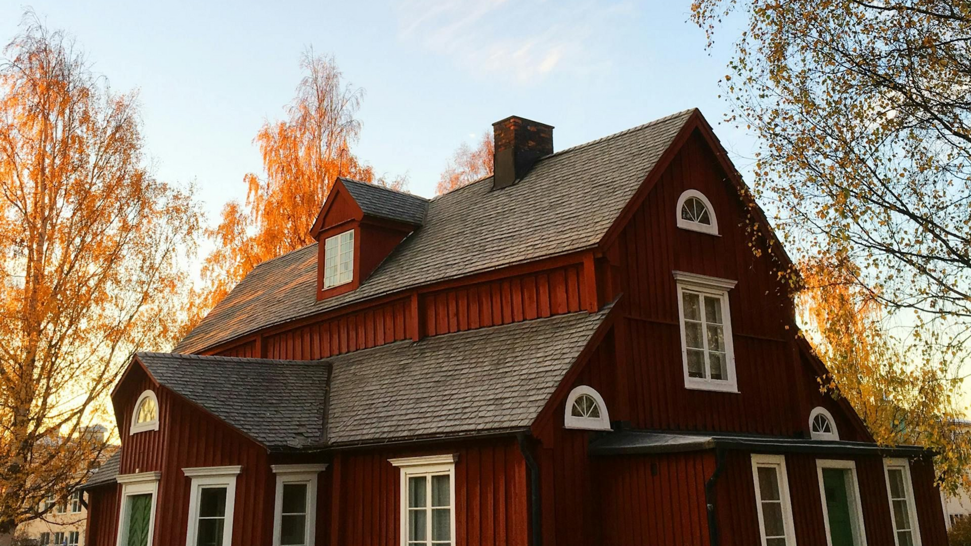 Red wooden building with white-framed windows under a blue and cloudy sky, surrounded by trees.