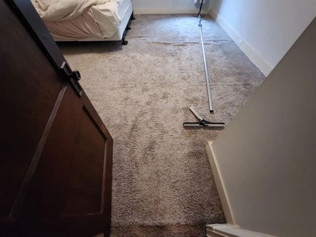A bedroom doorway reveals a light-colored carpet with a long-handled squeegee leaning on it.