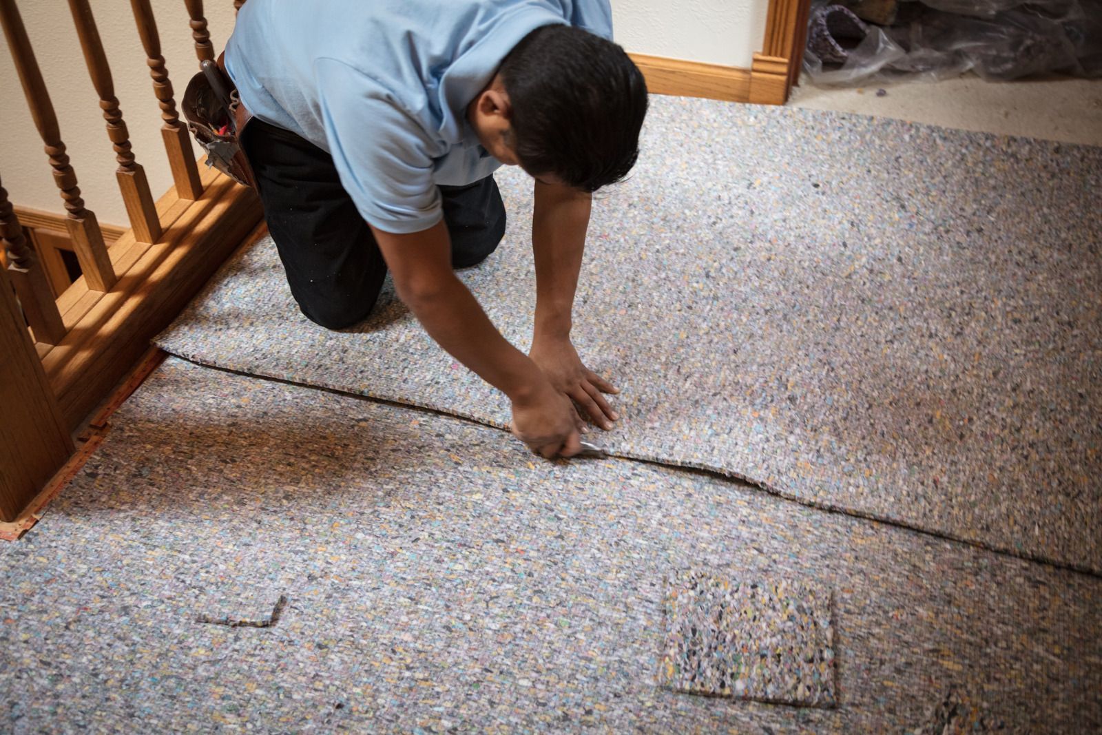 A person kneeling on a floor, using a utility knife to cut a section of gray speckled carpet padding.