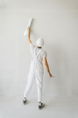 Person in white coveralls and hard hat smoothing plaster on a wall with a trowel.