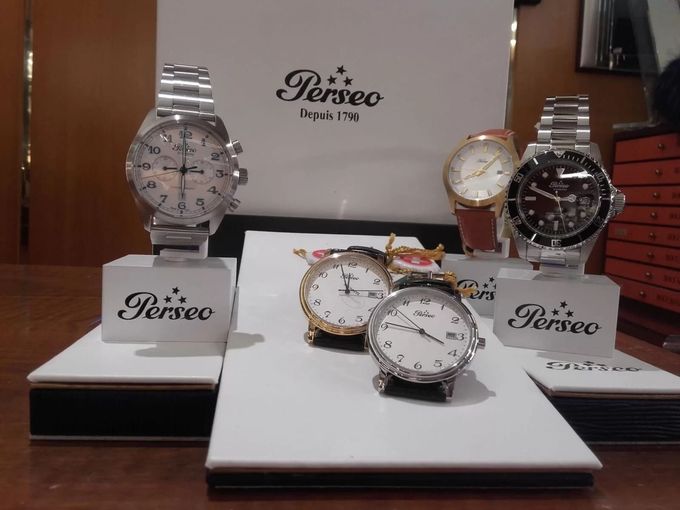 Esposizione di orologi Perseo su piattaforme bianche e nere. Include logo, cinturini in metallo e vari quadranti.