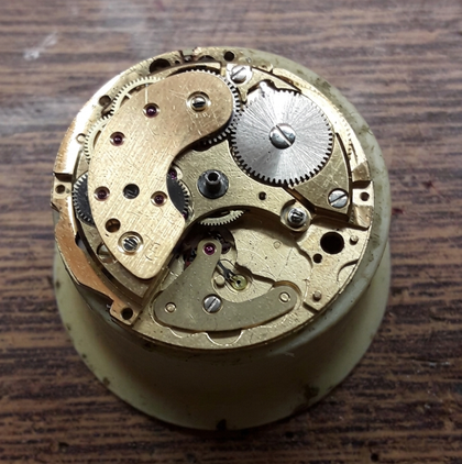 Meccanismo dell'orologio con vari ingranaggi, tonalità dorata, piccoli rubini, su base beige, superficie in legno.