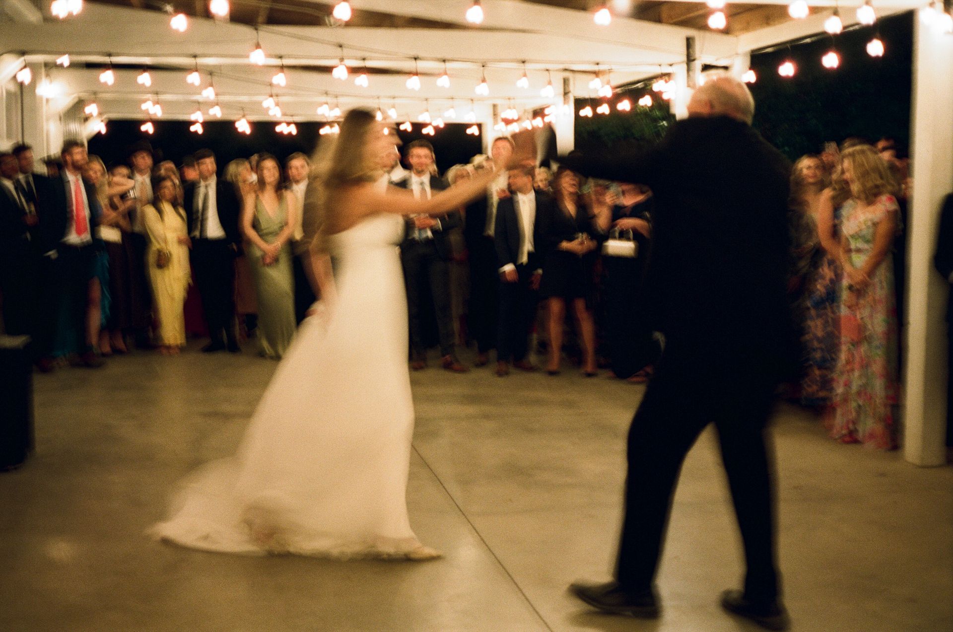 A bride in a white wedding gown dances with a partner in a dark suit under string lights at an evening reception.