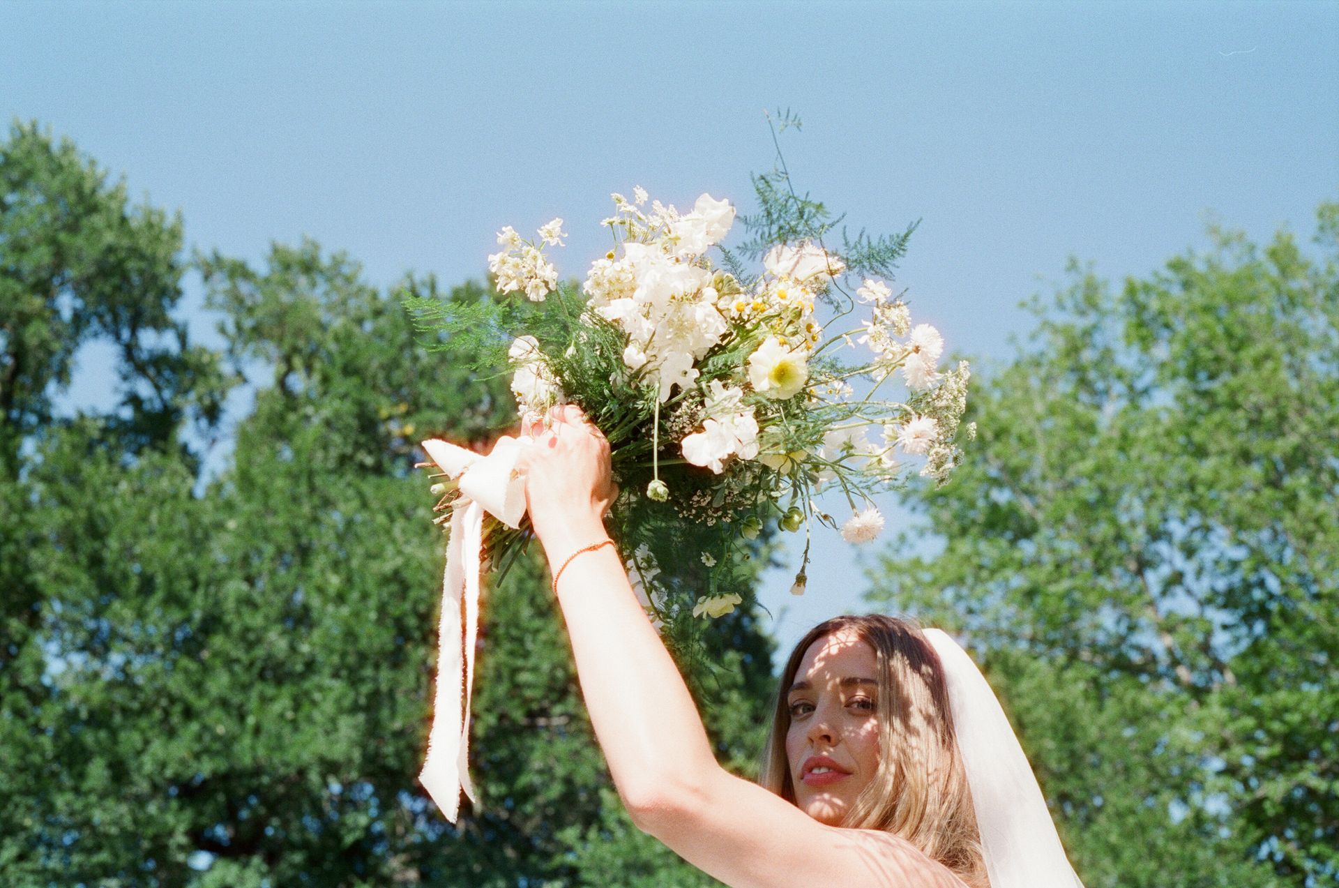 A person in a veil holds a white floral bouquet high toward a bright blue sky with trees in the background.
