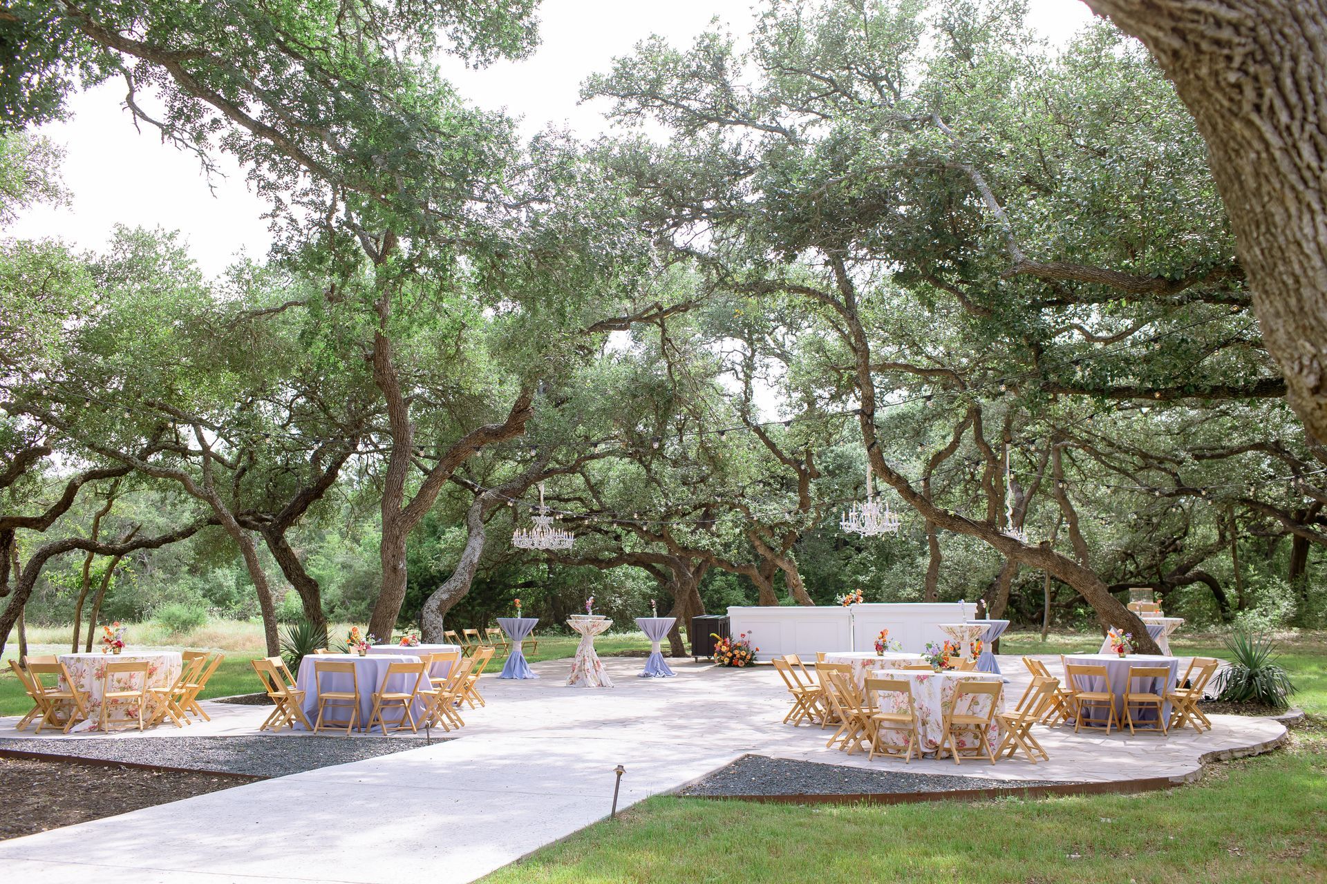 An outdoor event space with tables, chairs, and a bar set up on a stone patio beneath a canopy of large oak trees.