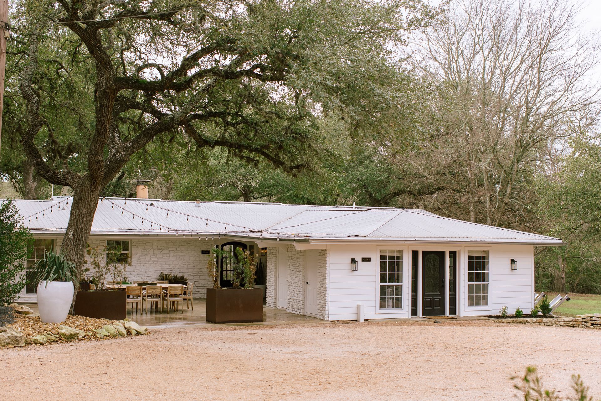 A white, single-story house with a metal roof and a gravel driveway, framed by large trees and a patio area.