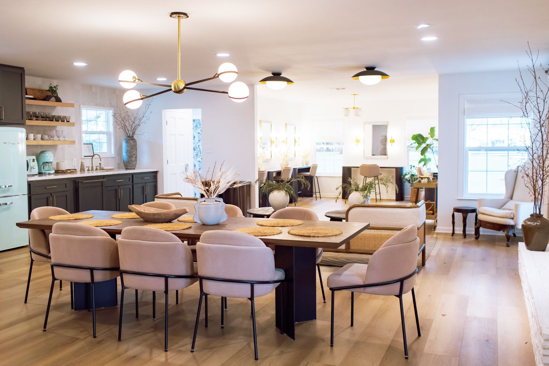 Dining room with a long table, six light-pink upholstered chairs, and a modern chandelier, adjacent to a kitchen.