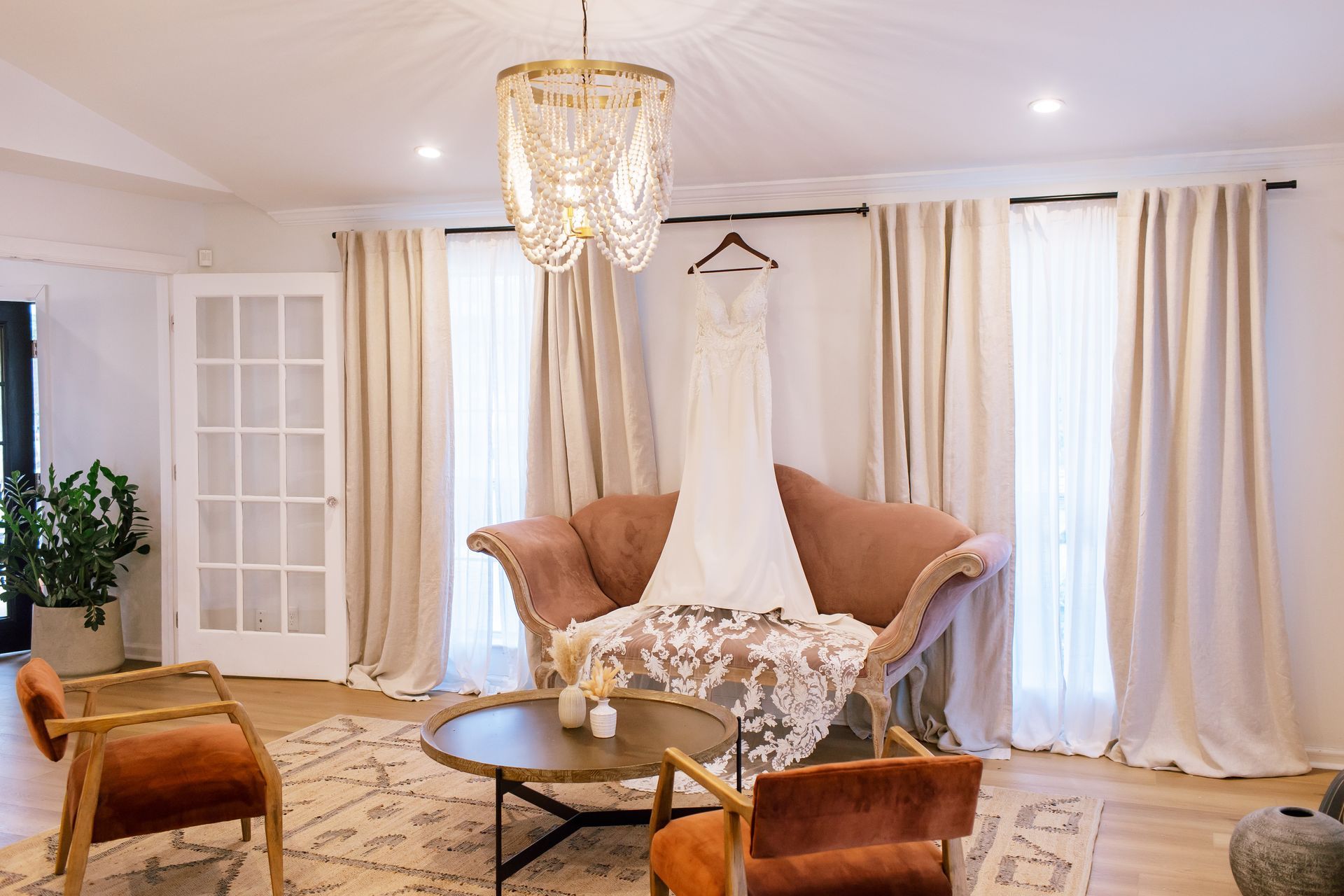 A white wedding dress hangs above a pink velvet settee in a bright, modern room with neutral decor and a central chandelier.