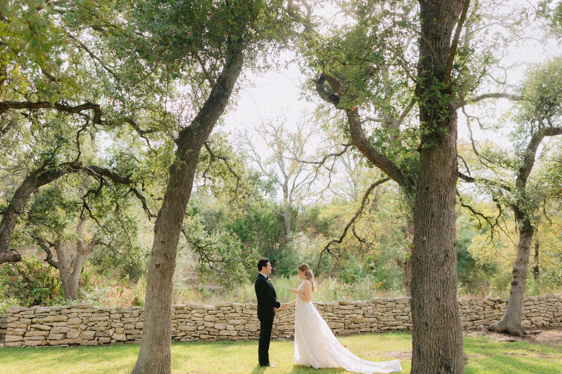 A couple stands facing each other in a grassy area framed by trees, in front of a low stone wall.