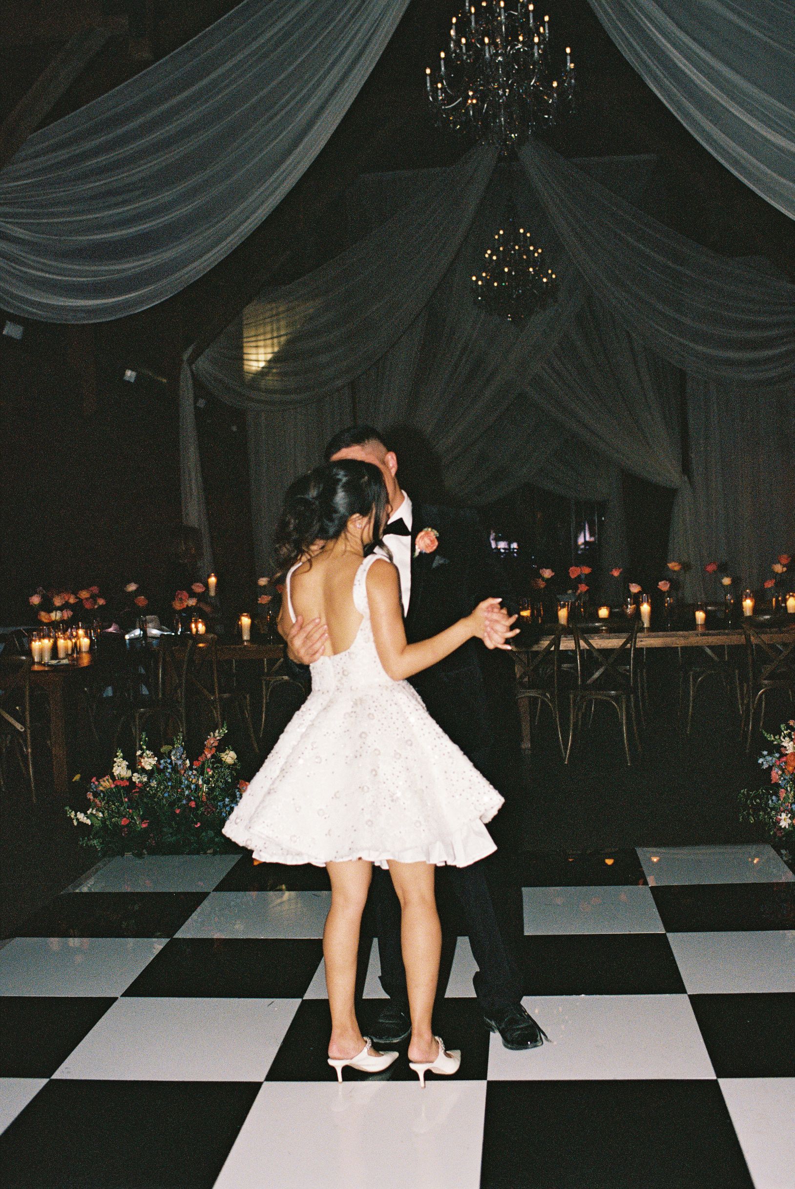 A couple in formal attire dances on a black-and-white checkered floor in a dimly lit, decorated ballroom.