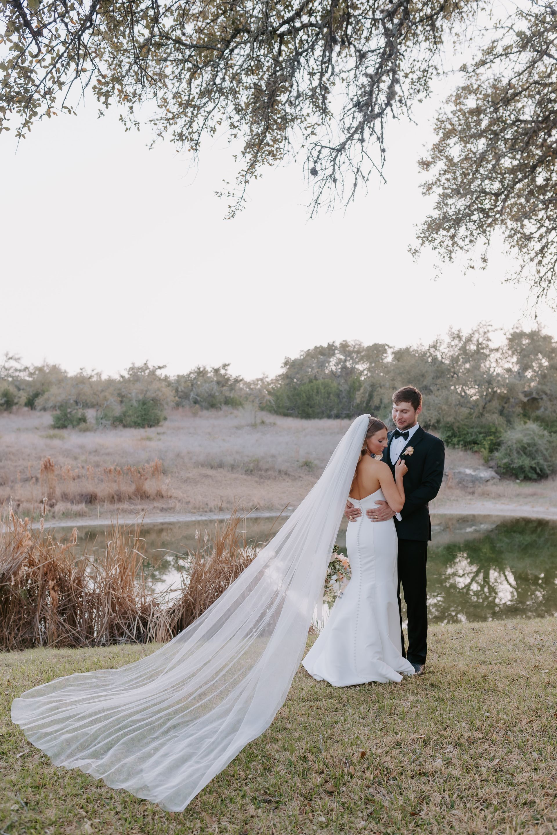 A bride and groom standing beside a pond under trees, with the bride's long, flowing veil trailing on the grass.