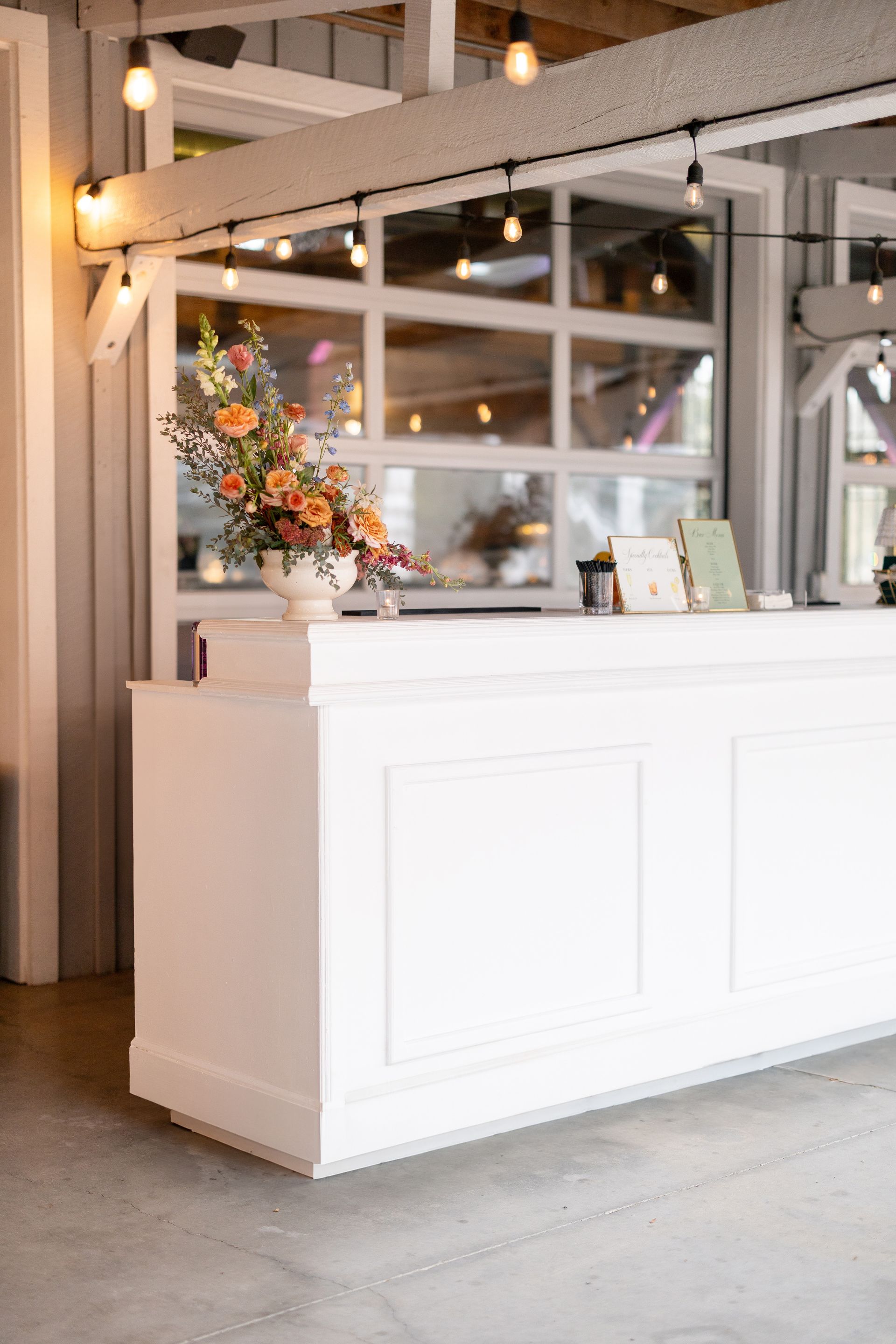 A long white reception bar with a floral centerpiece under hanging Edison lights in a rustic venue.