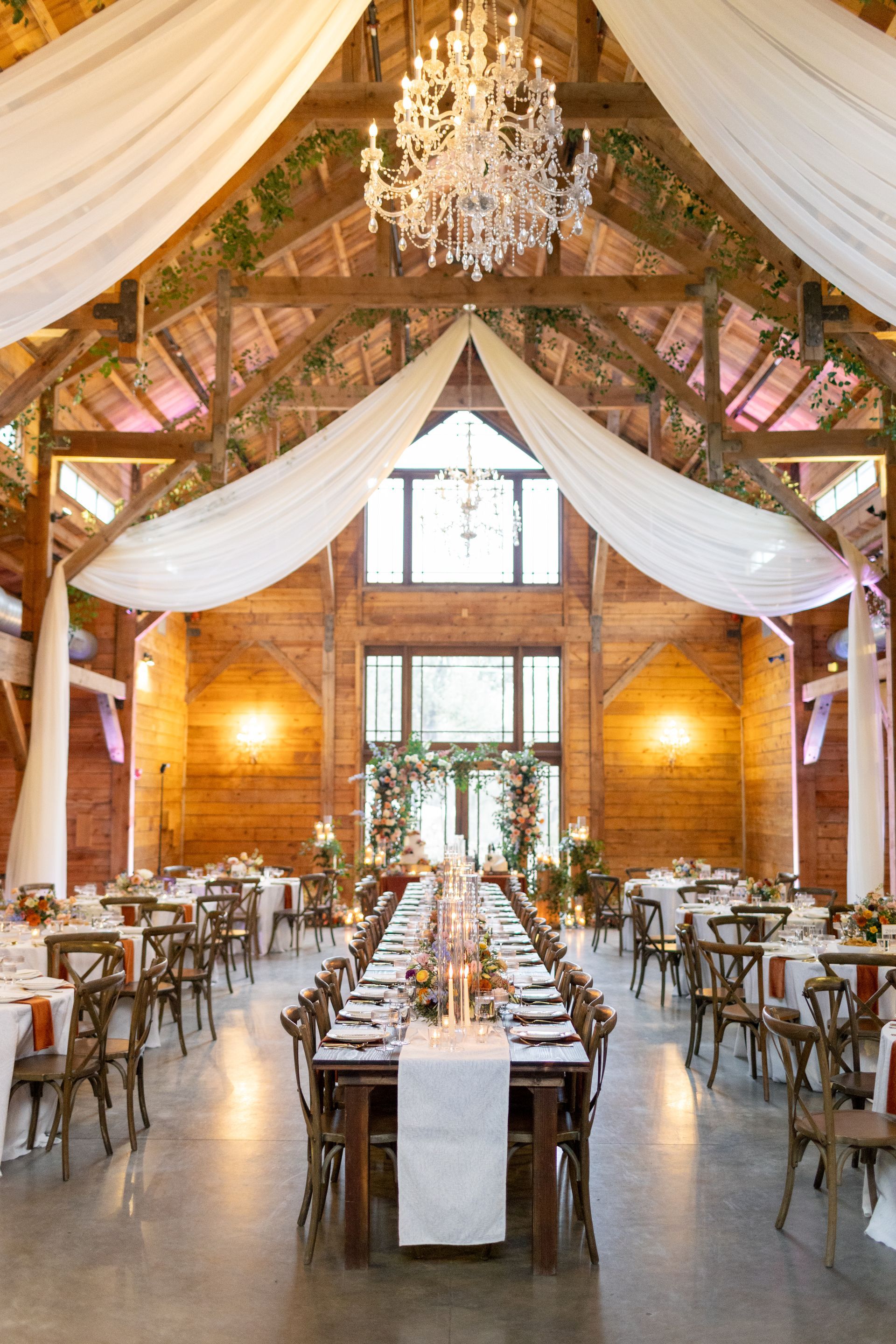 Rustic barn wedding venue interior with draped white fabric, wood beams, a crystal chandelier, and long dining tables.