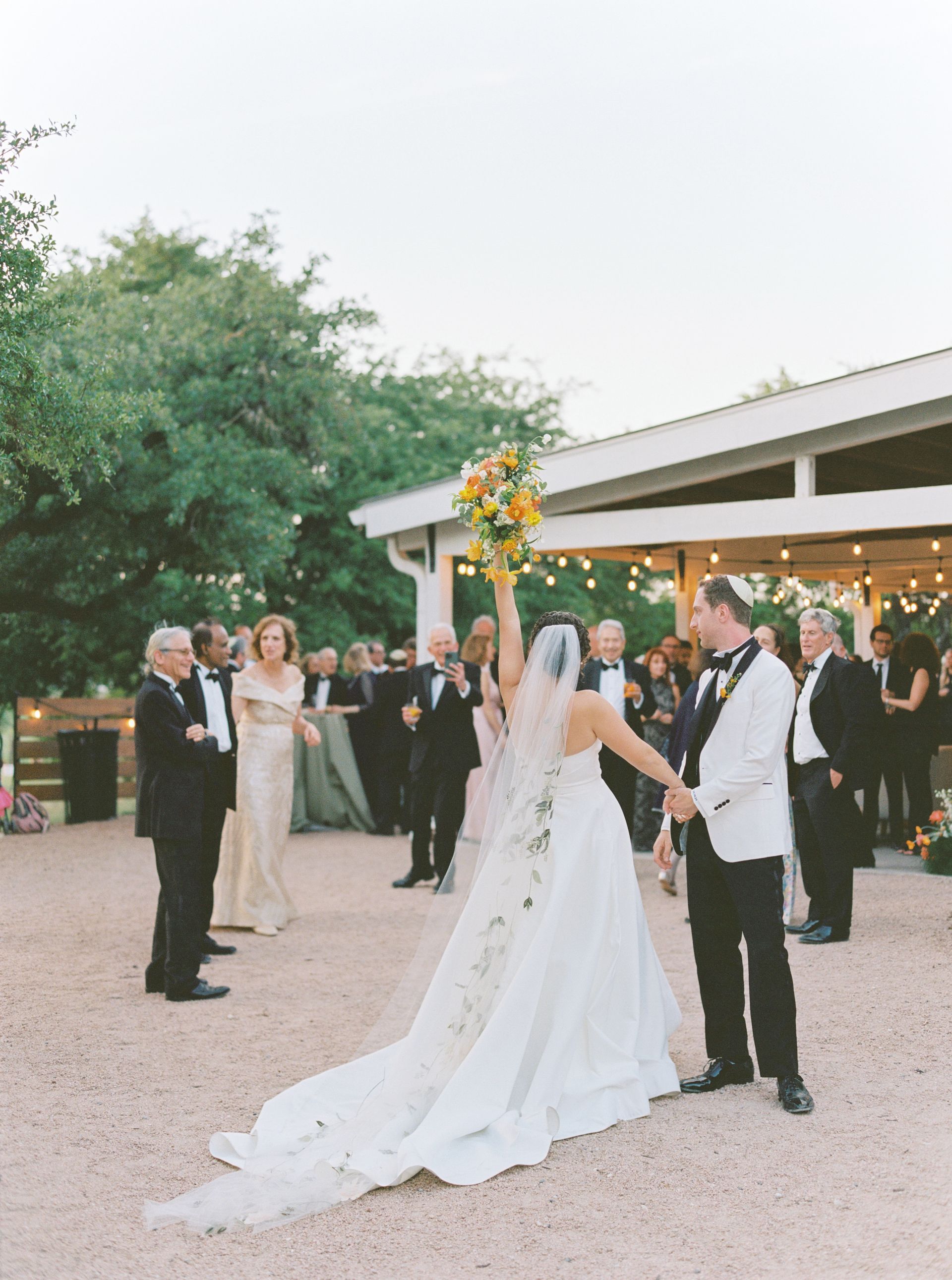 A bride holds her bouquet aloft while holding hands with a groom on a gravel path in front of a wedding reception.