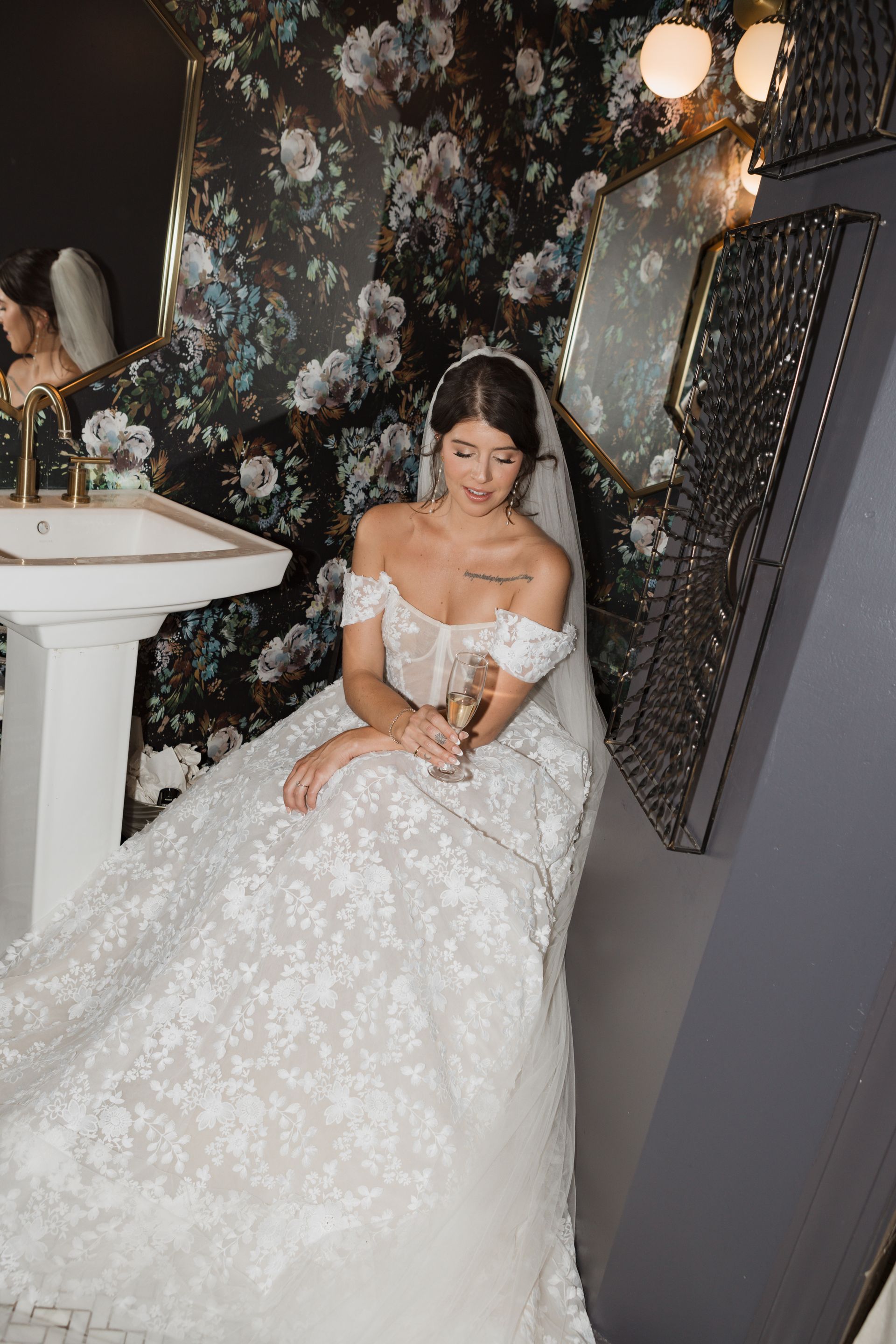 A person in a lacy, off-the-shoulder wedding gown and veil sits holding a glass in a dark, floral-wallpapered restroom.