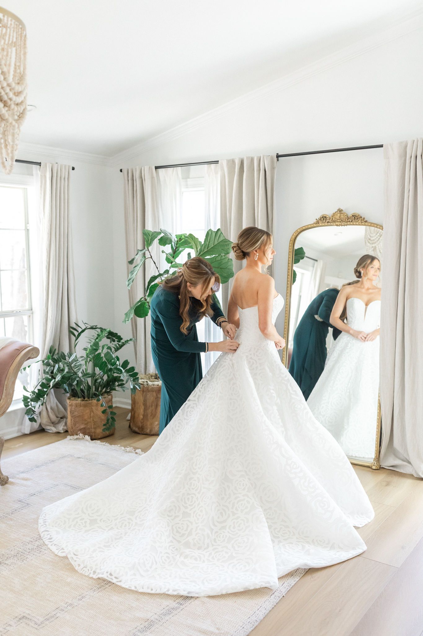 A person in a dark top assists a bride in a voluminous white wedding dress with a long train in a bright, modern room.