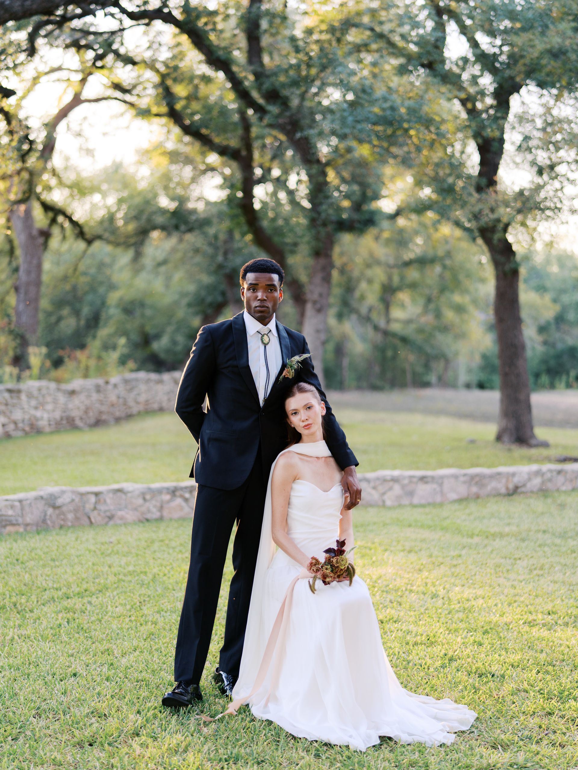 A couple poses for a formal portrait in a grassy meadow with stone walls and trees in the background at sunset.