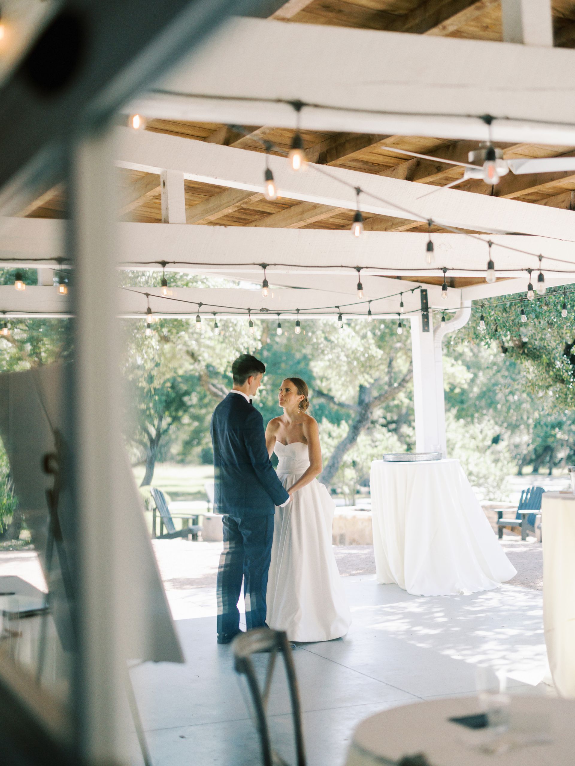 A couple faces each other in a wedding embrace under a wooden patio structure decorated with string lights.